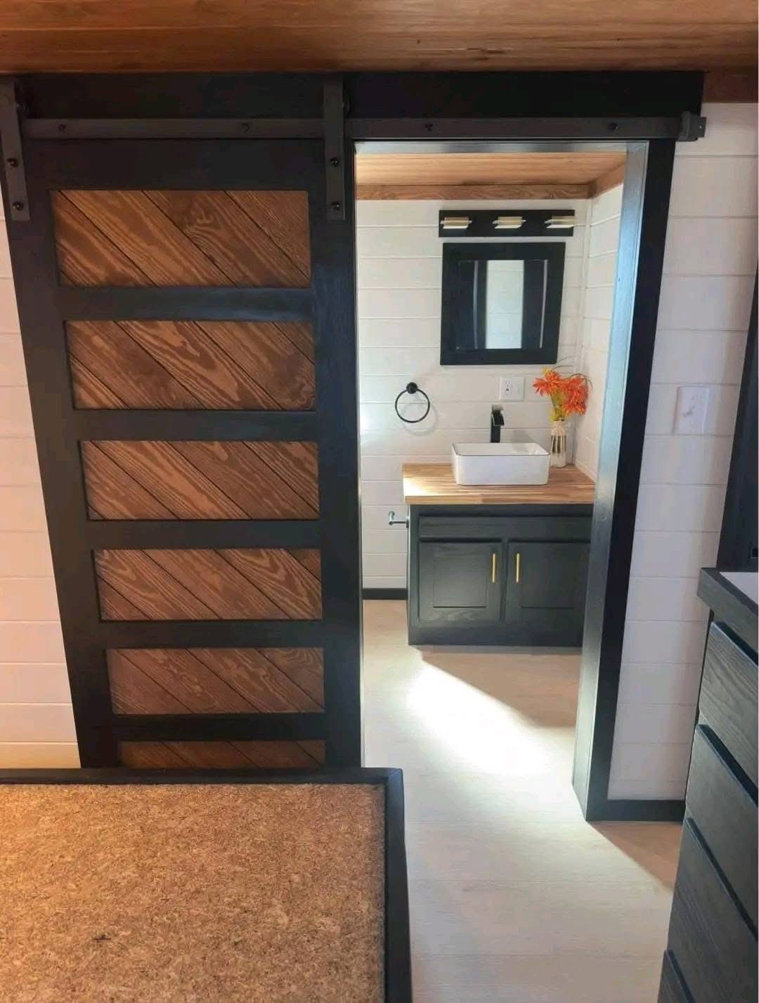 Modern bathroom with a rustic sliding barn door. Inside, black vanity with a white vessel sink, a mirror, and orange flowers conveys warmth and elegance.