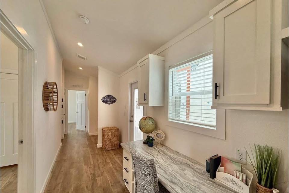Bright hallway with wood flooring, white walls, and ceiling-lit fixtures. A console table holds a globe and decor items under a window with blinds. Calm ambiance.