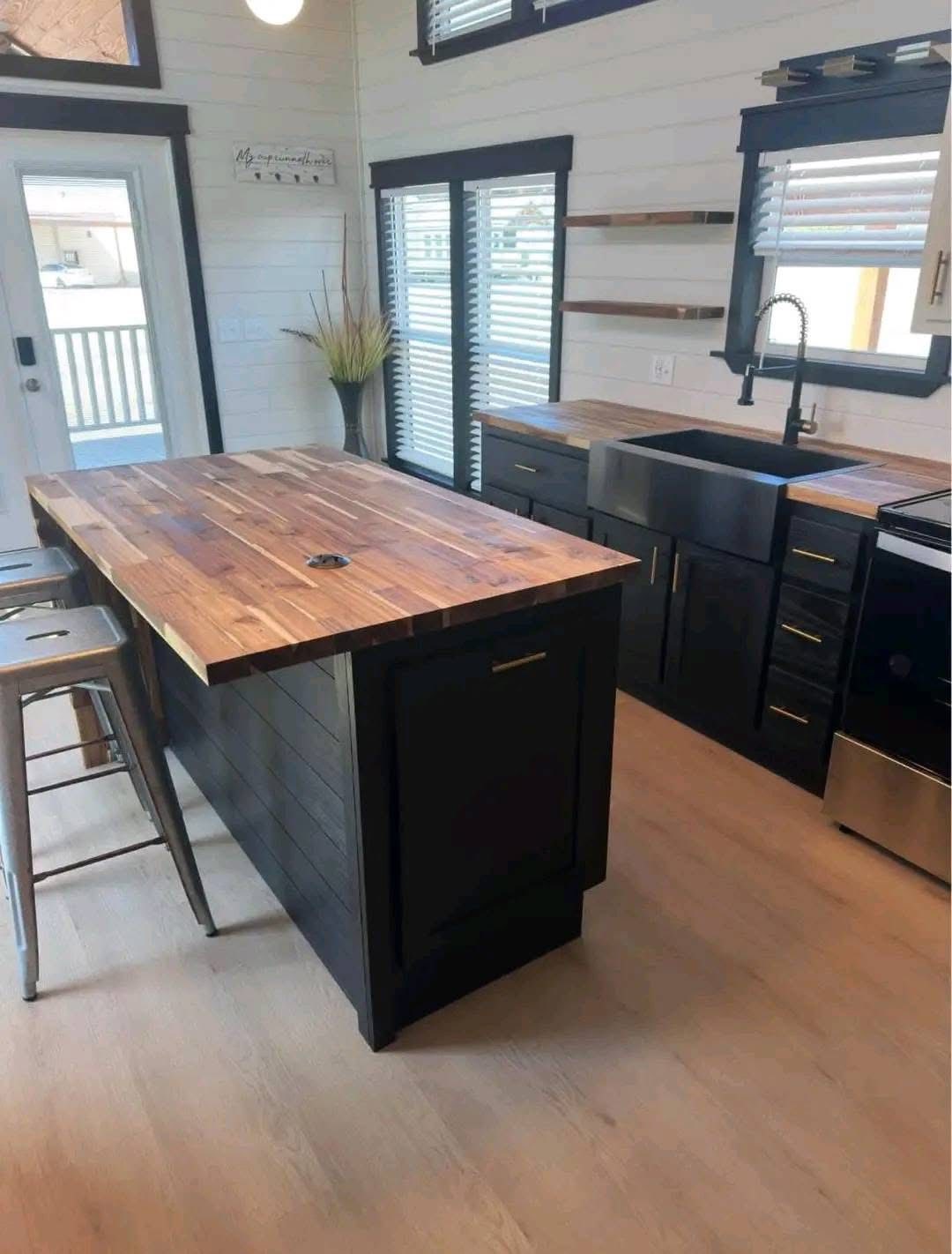 Modern kitchen with a sleek black island and wooden countertop, surrounded by black cabinets. Light wood floors and large windows add brightness.