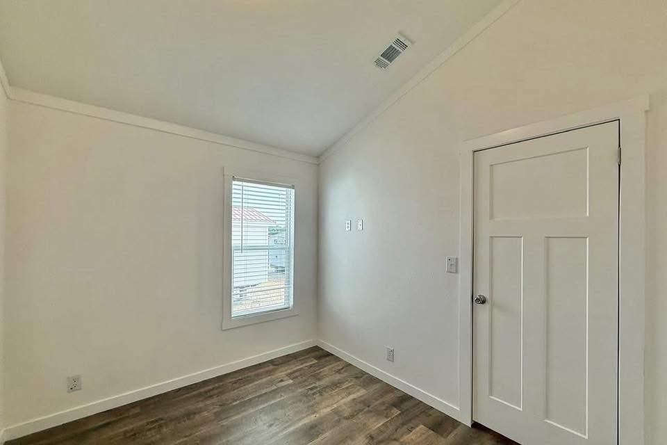 A small, empty room with white walls and a wooden floor features a single window with blinds, a closed white door, and a sloped ceiling, evoking a minimalist vibe.