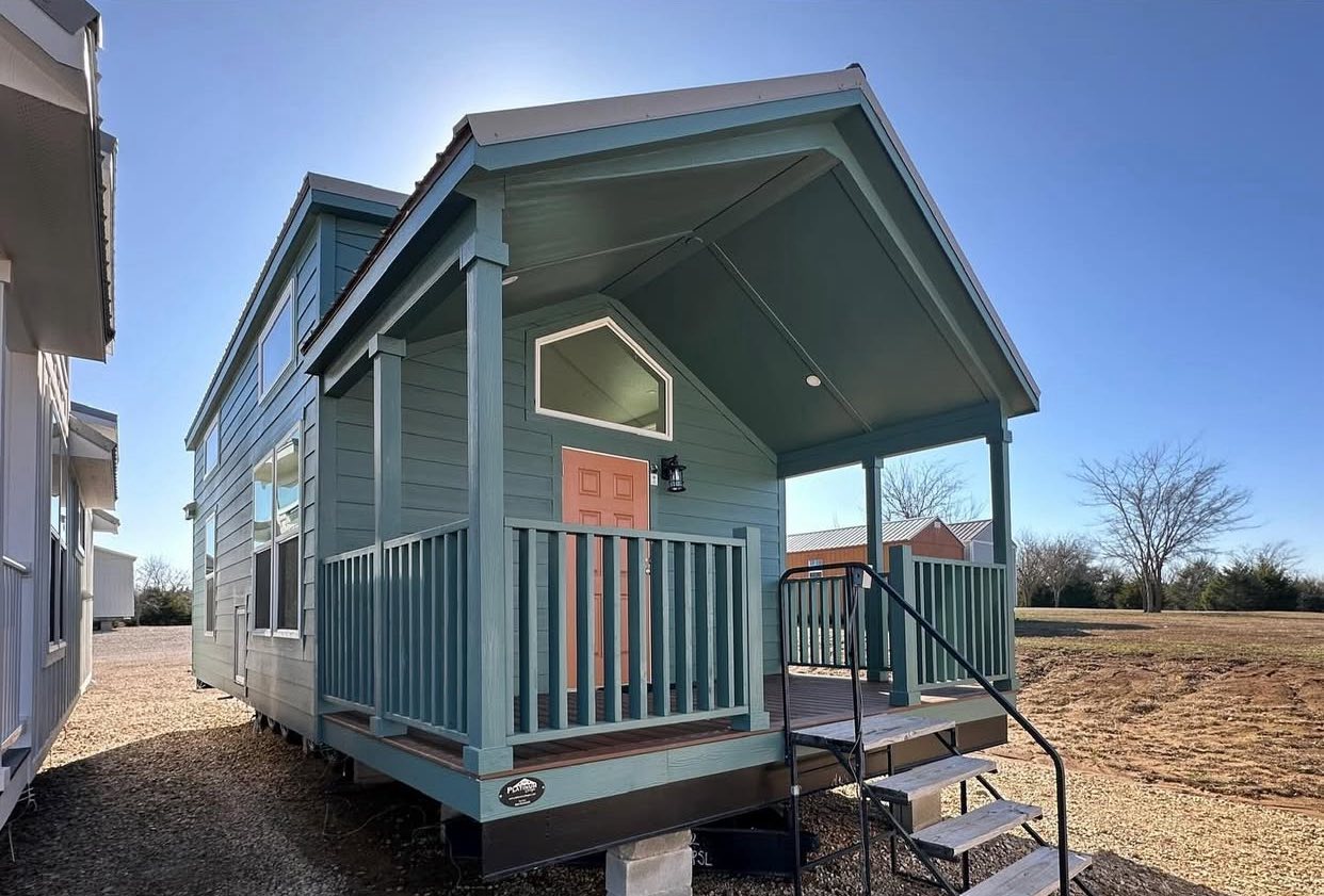 A blue tiny house with a gabled roof and porch sits against a clear sky. The coral door adds a pop of color. Steps lead up to the porch, conveying coziness.