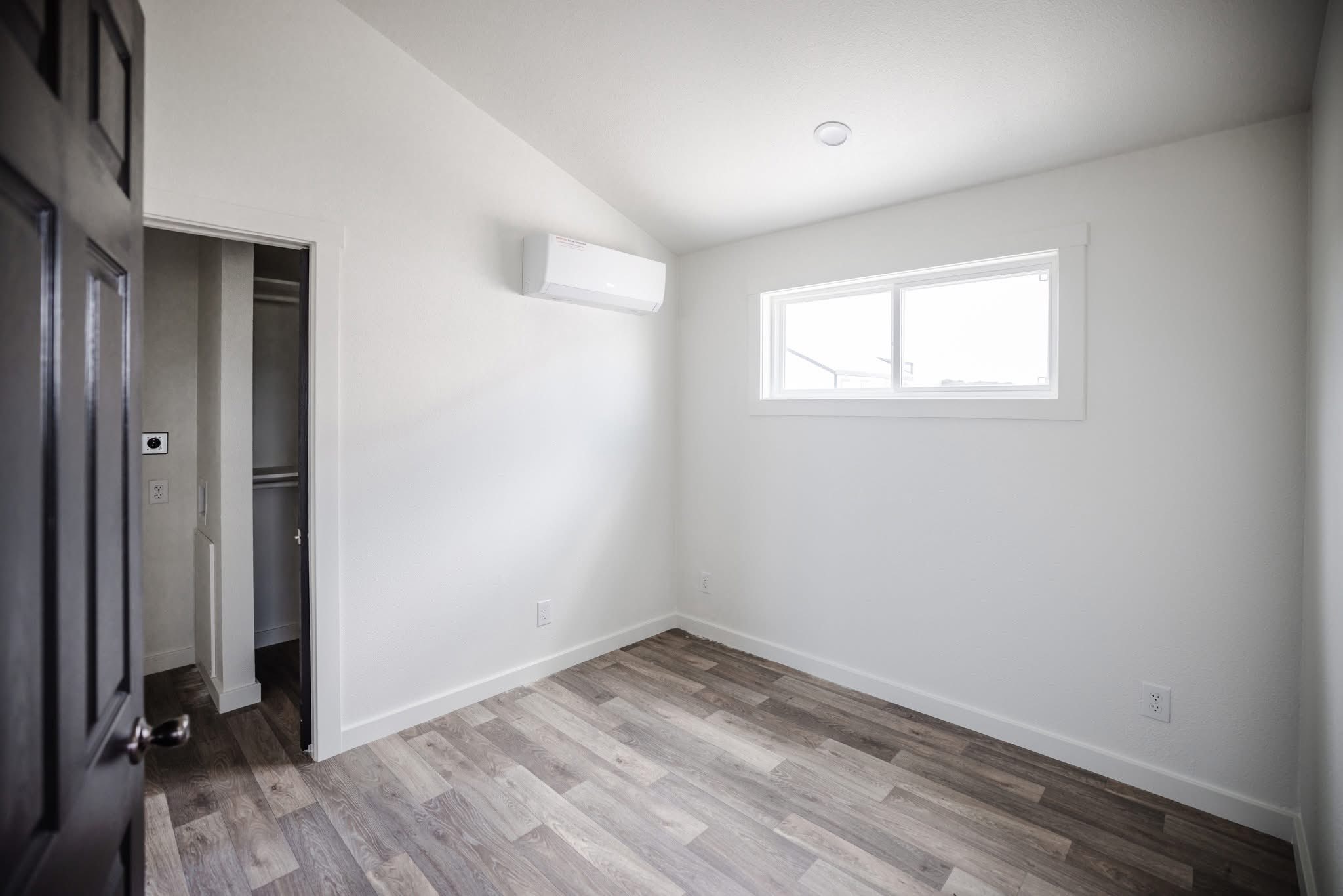 Minimalist room with light wood flooring, white walls, and a small rectangular window. An air vent is mounted high on one wall. The room is empty, conveying simplicity and openness.