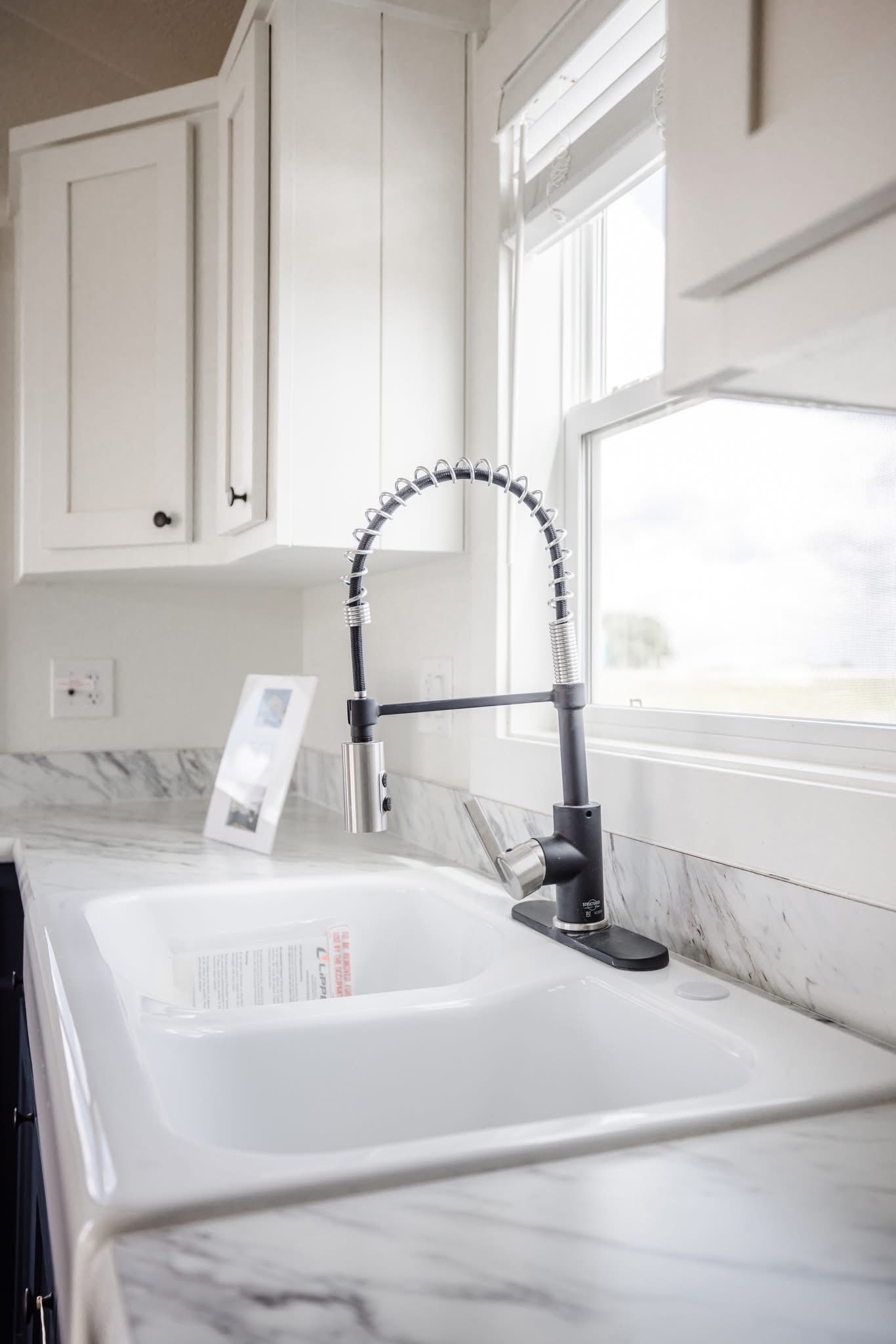 Close-up of a modern kitchen sink with a sleek, high-arched faucet, set against white cabinets and a marble countertop. Sunlight filters through a window.