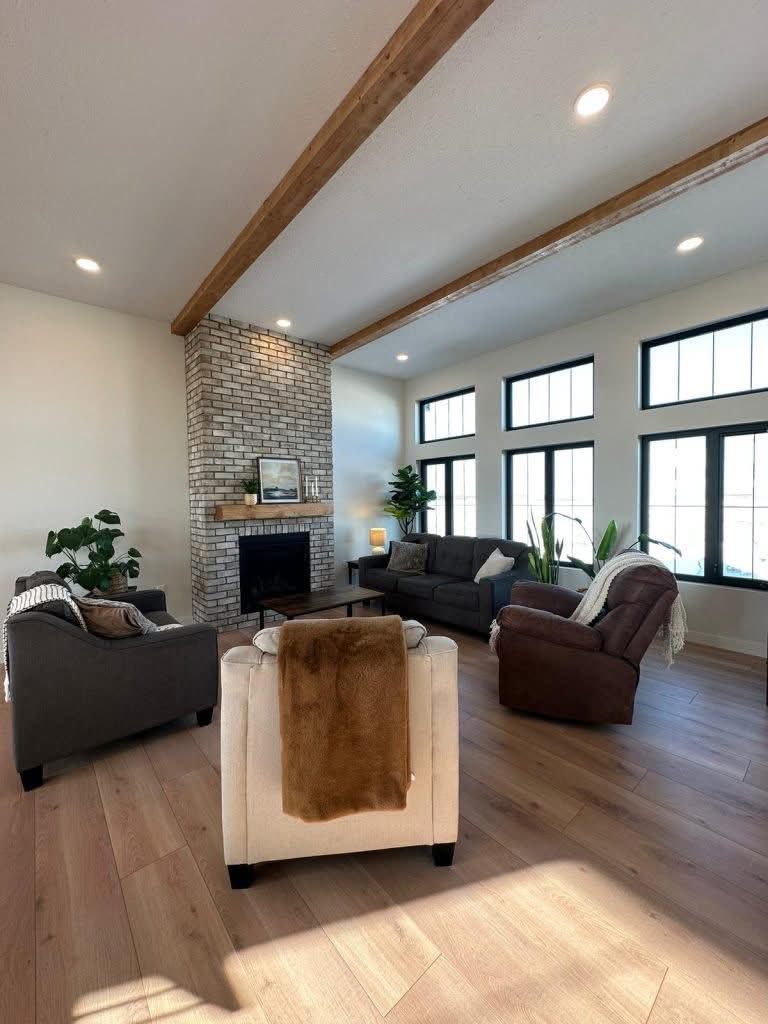 Spacious living room with brick fireplace, wooden beams, large windows, and cozy seating. Minimalist decor with plants, creating a warm, inviting atmosphere.