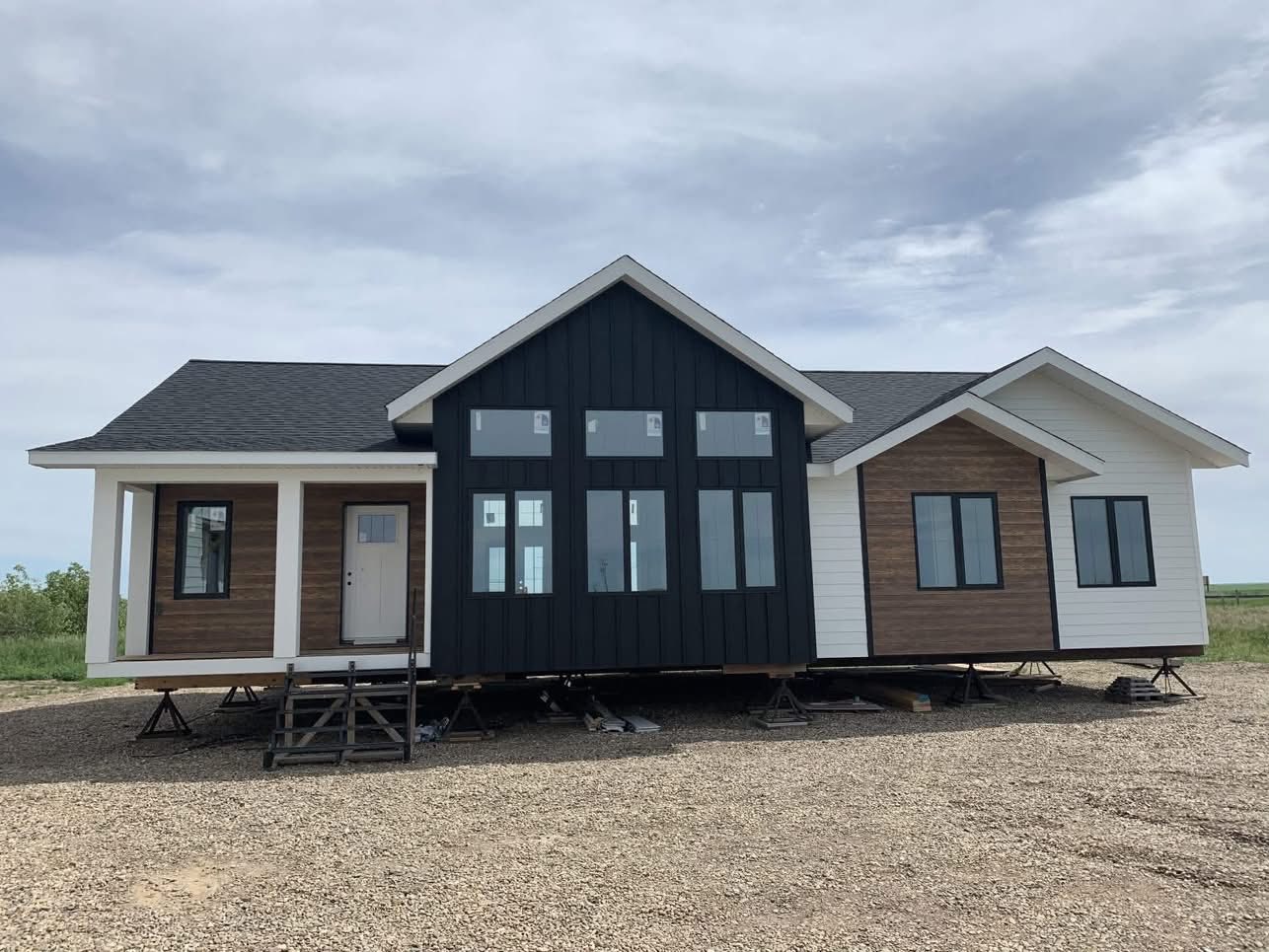 Modern modular home on gravel, elevated on supports. Features a mix of dark and light siding, large windows, and a cloudy sky in the background.
