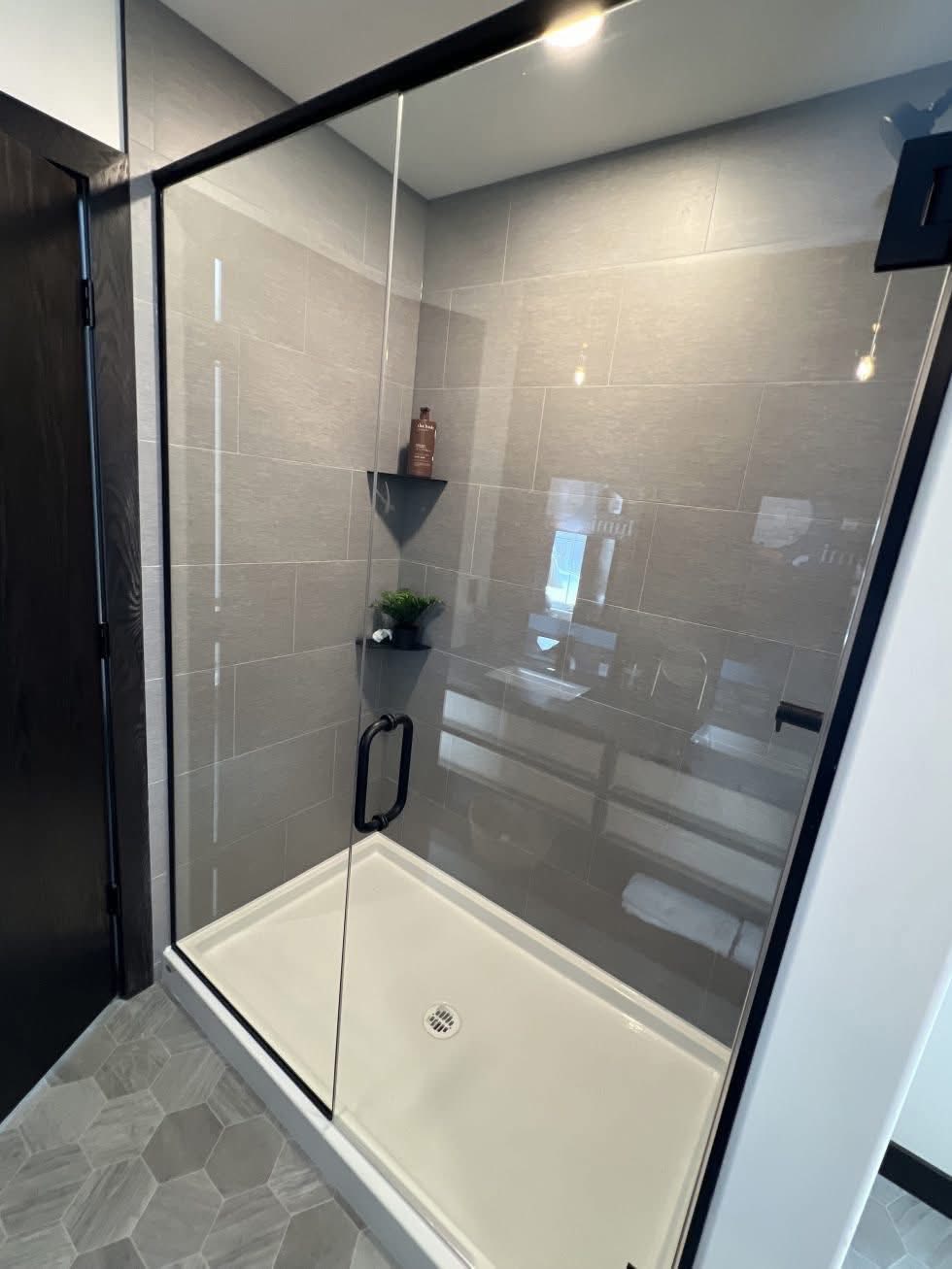 Modern bathroom shower with glass door, gray tiles, and hexagonal floor tiles. Inside are a brown bottle and a small plant on corner shelves.