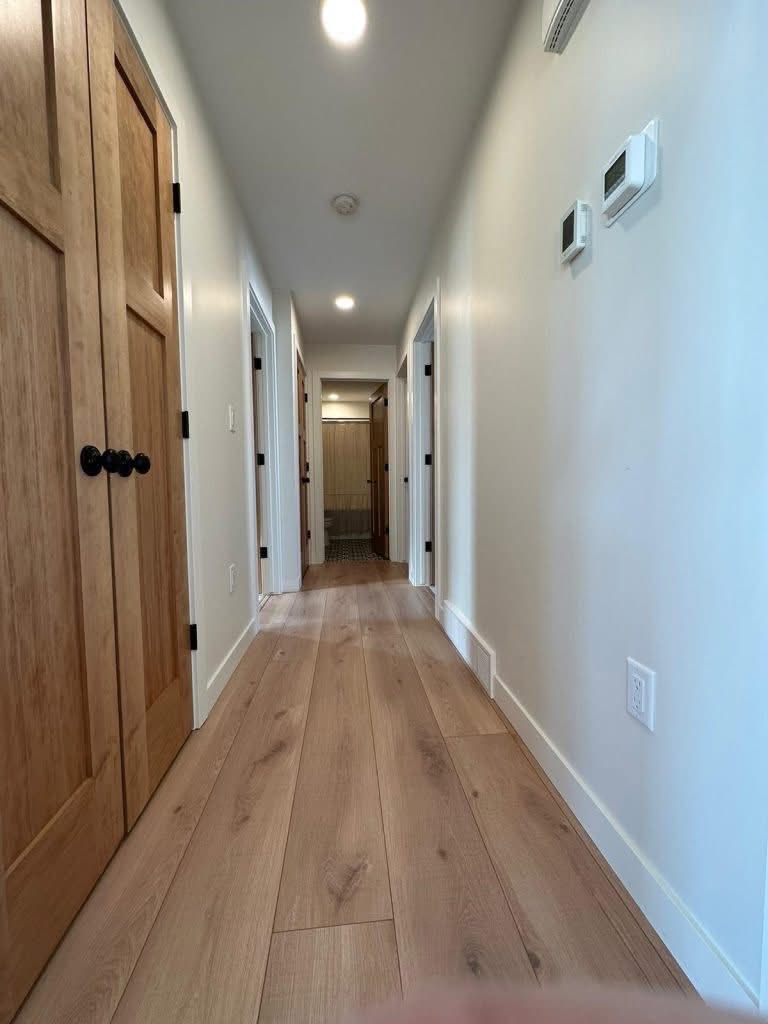 A long, narrow hallway with light wooden flooring and white walls. Wooden doors with black handles line the sides. Ceiling lights create a cozy atmosphere.