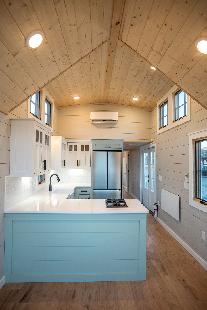 Small, modern kitchen with light wood paneling, white cabinets, stainless steel fridge, light blue island, and warm wood floors, creating a cozy feel.