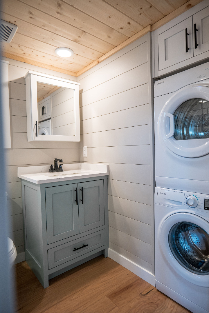 Compact laundry room with wood ceiling and floor. Features a stacked washer and dryer beside a light gray vanity with a mirror above. Bright and cozy.