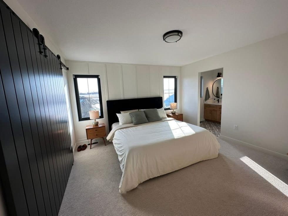 A modern bedroom with a white and black theme features a large bed with a dark headboard, flanked by wooden nightstands. Two black-framed windows allow natural light in.