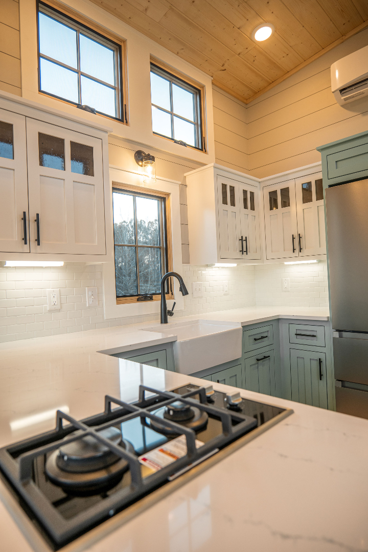 Bright kitchen with white cabinets, black handles, and a gas stove on a marble countertop. Large windows and wood ceiling add warmth. Modern and inviting.
