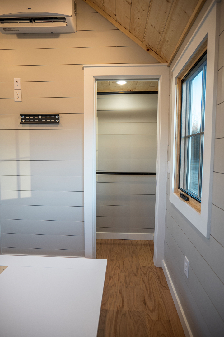 Small interior with wood-paneled walls, open closet, and light wood floor. Features a window, air conditioner, and TV mount, creating a cozy atmosphere.