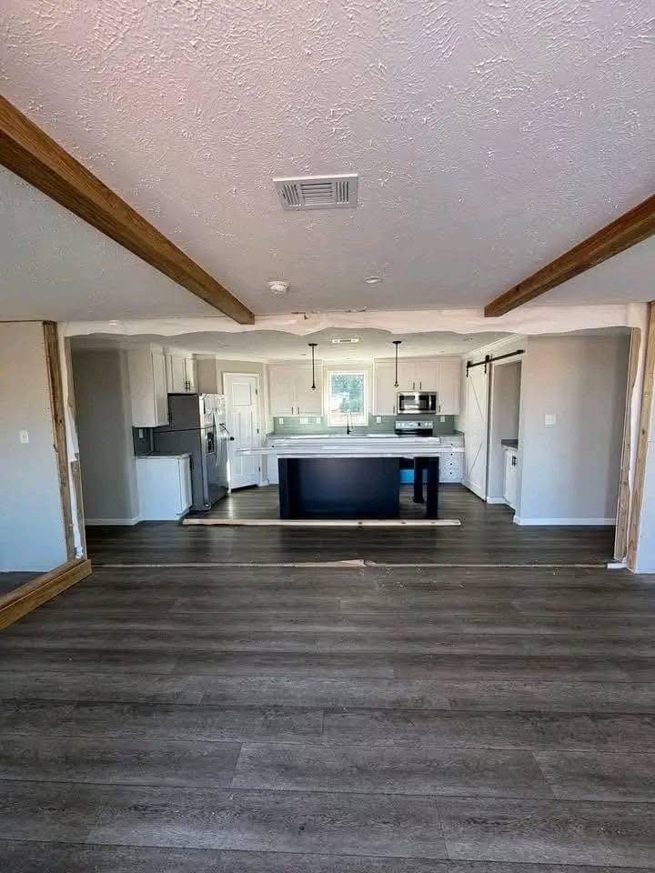Modern kitchen with light gray cabinets, dark island, and stainless steel appliances. Wooden ceiling beams and dark laminate flooring add warmth.
