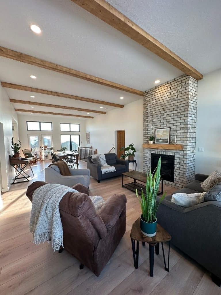 Spacious living room with wooden beams, a brick fireplace, cozy seating, and potted plants. Large windows provide natural light, creating a warm atmosphere.