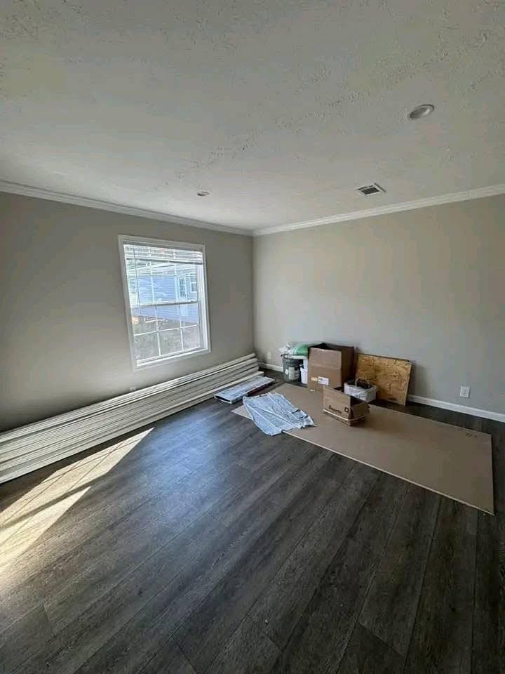 Empty room with light gray walls and dark wood flooring. Sunlight streams through a window. Materials and boxes are stacked in the corner, suggesting renovation.