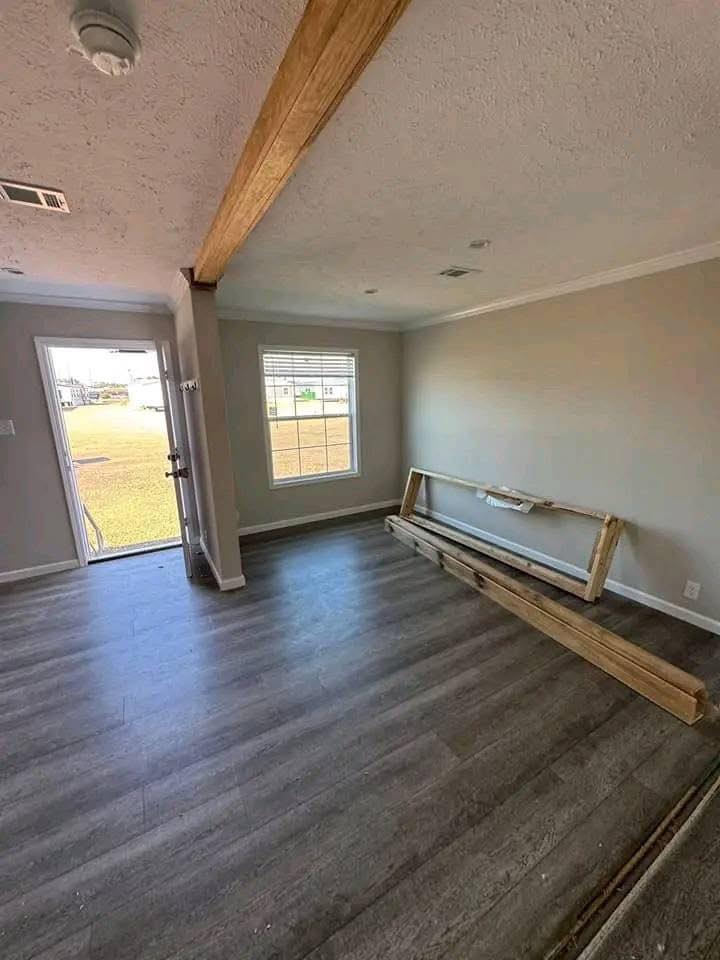Empty room with gray wood floors, white walls, and a wooden beam on the ceiling. A window and open door let in light; lumber rests against a wall.