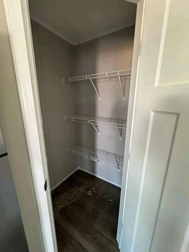 A small, empty closet with two white wire shelves against a gray wall. The wooden floor shows debris in the corner, suggesting a need for cleaning.