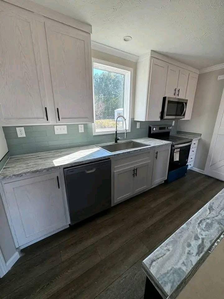 Modern kitchen with white cabinets, marble countertops, and stainless steel appliances. Sunlit window above sink and wood flooring enhance warmth.