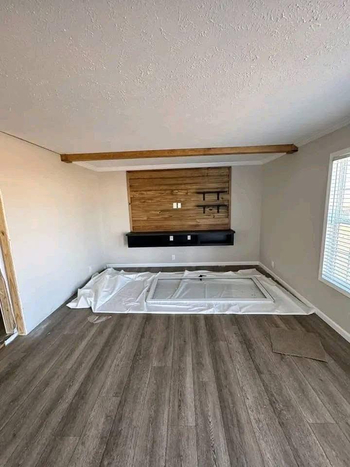 A room with light gray walls, wood flooring, and a large window on the right. A wooden accent wall with a dark shelf and small shelves is visible.