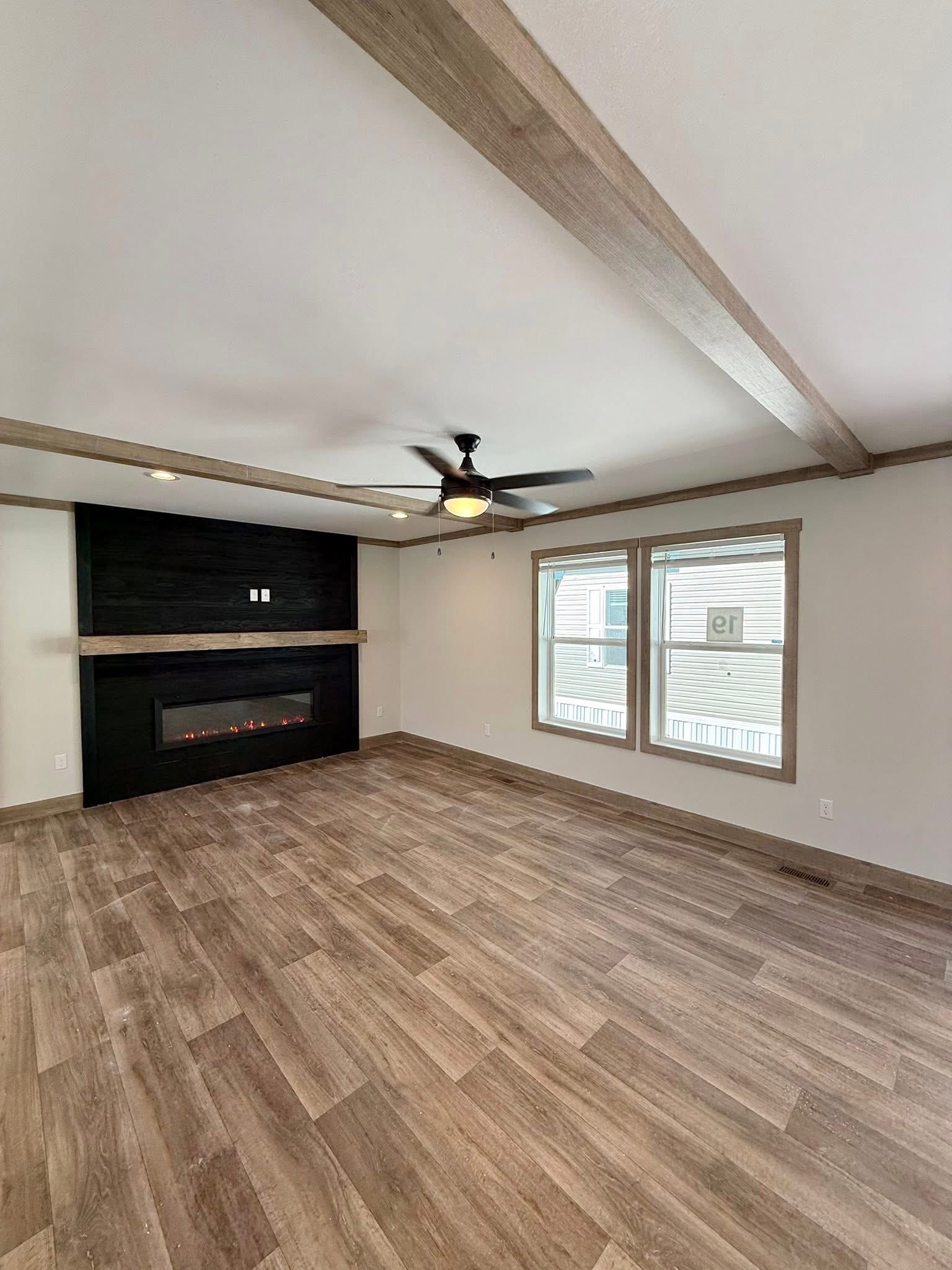 Spacious living room with wooden floors, exposed beams, and a sleek black fireplace. A ceiling fan and two large windows add light and warmth.