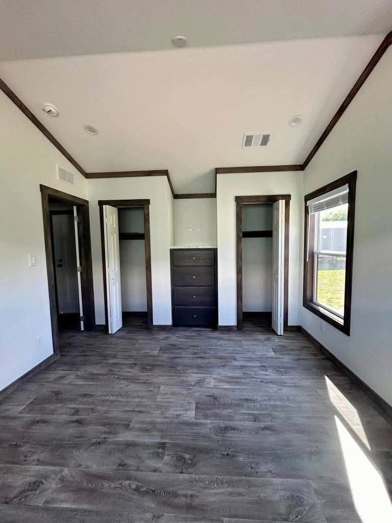 A bright, empty room with wood floors, white walls, and dark-trimmed closets. Natural light enters through a large window, creating a calm, open feel.