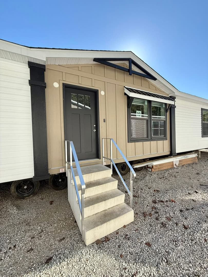 A tan and white modular home with a pitched roof, black door, and twin windows. Concrete steps with metal railings lead to the entrance. Bright, sunny day.