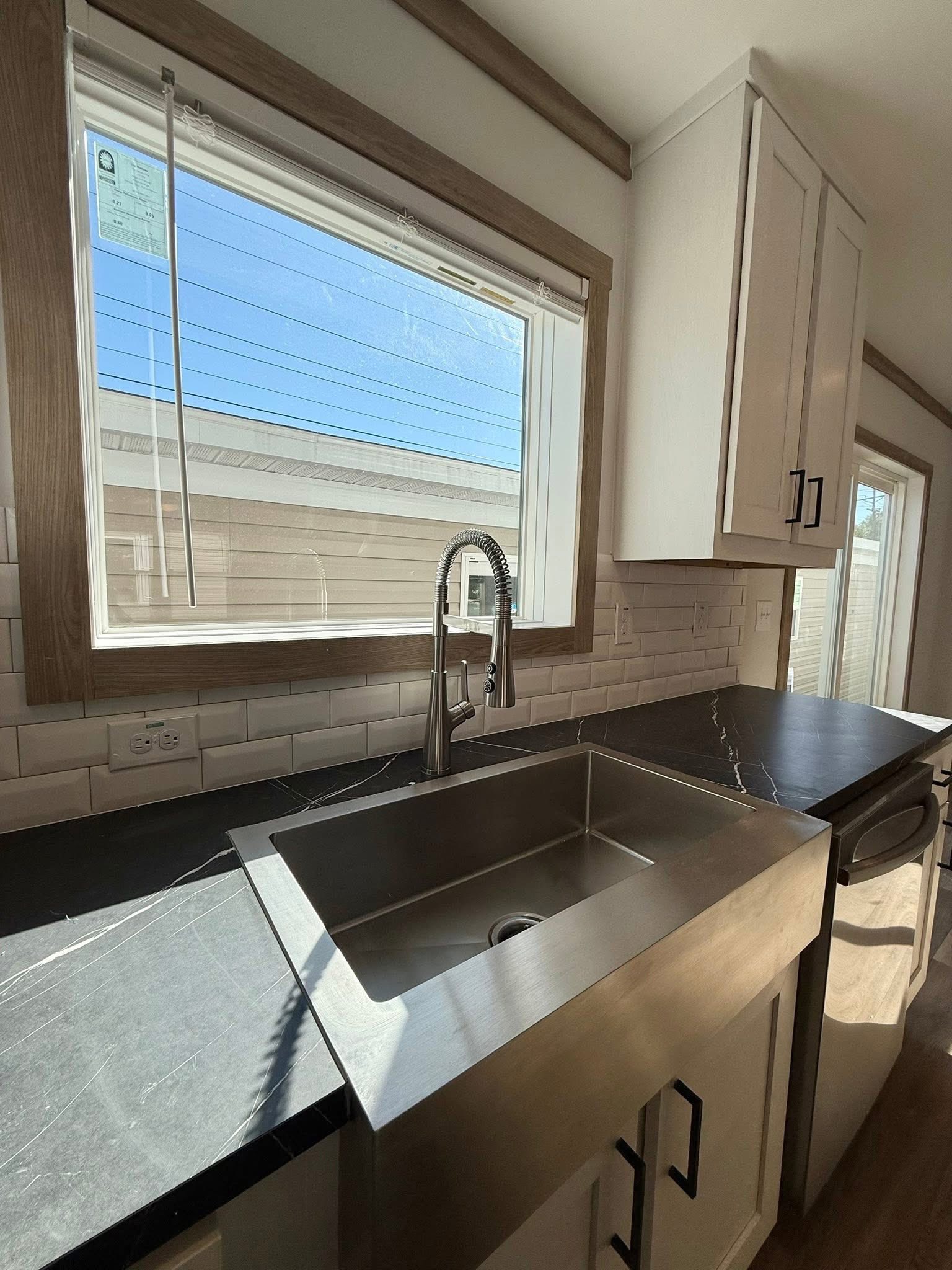 A modern kitchen with stainless steel sink and faucet, dark marble countertop, and white cabinets. Sunlight streams through a large window, creating a bright and clean atmosphere.