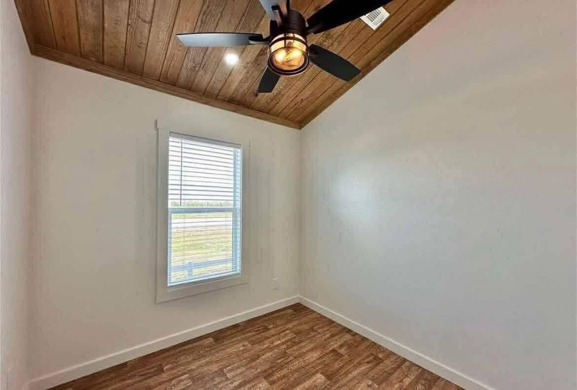 Small, empty room with white walls and a wooden floor. A sloped wooden ceiling features a modern black ceiling fan. Light streams through a window with blinds.