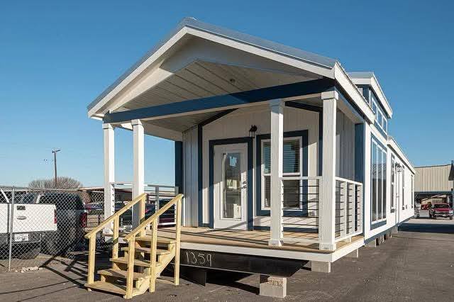 A modern tiny house with blue and white siding, featuring a covered porch and wooden steps, sits on a sunlit lot, evoking a cozy and inviting atmosphere.