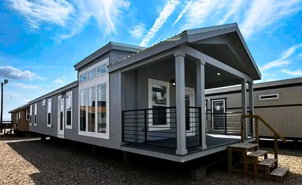 Modern gray modular home with large windows and a small porch, set in a gravel lot under a bright blue sky with wispy clouds.