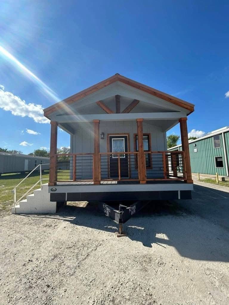 Cozy tiny house with a wooden porch, set against a clear blue sky. The structure is elevated, accessible by steps, conveying a welcoming atmosphere.