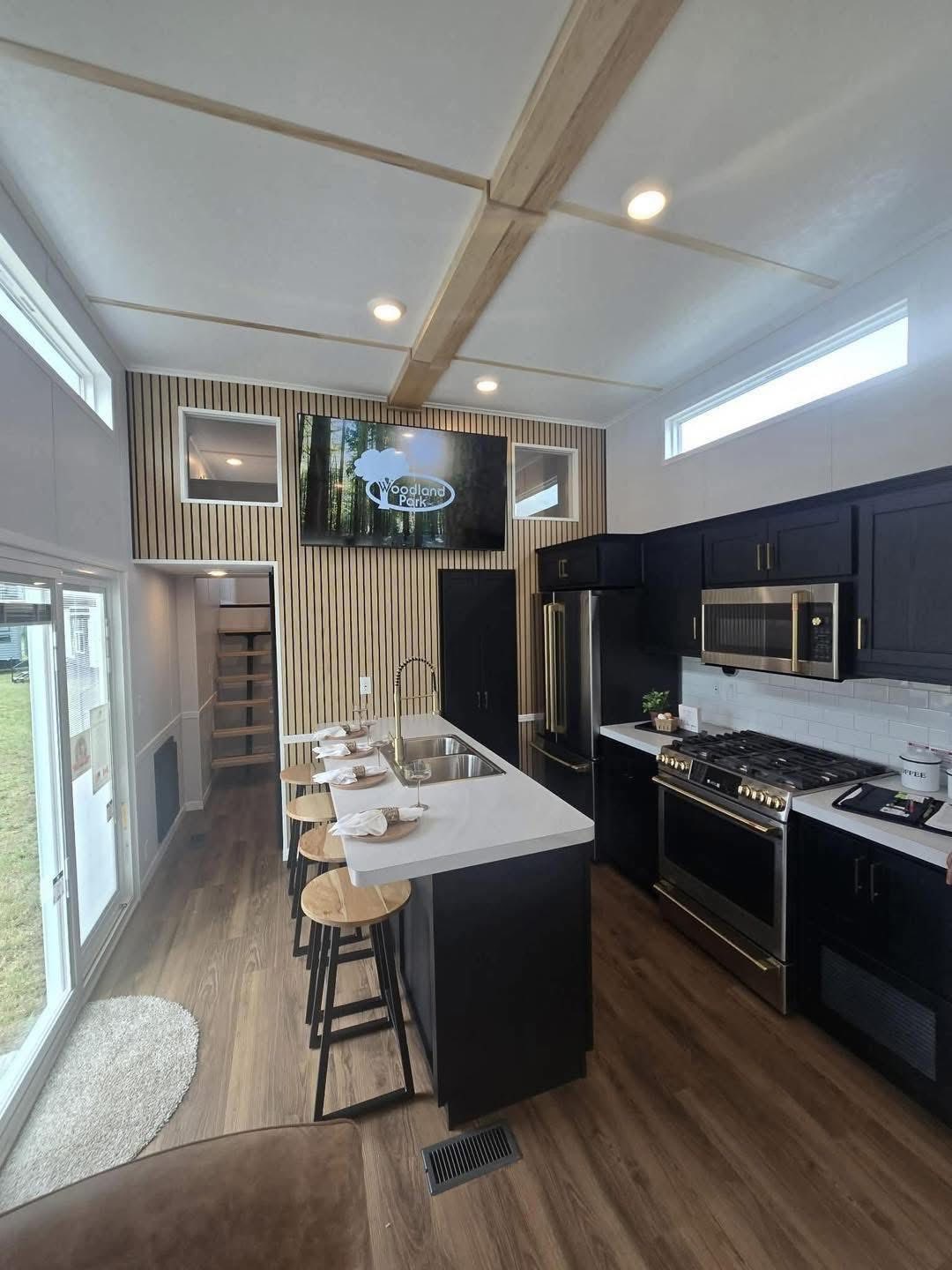 Modern kitchen with black cabinets, stainless steel appliances, and a center island with wooden stools. Bright lighting and a wall TV create a cozy, inviting atmosphere.