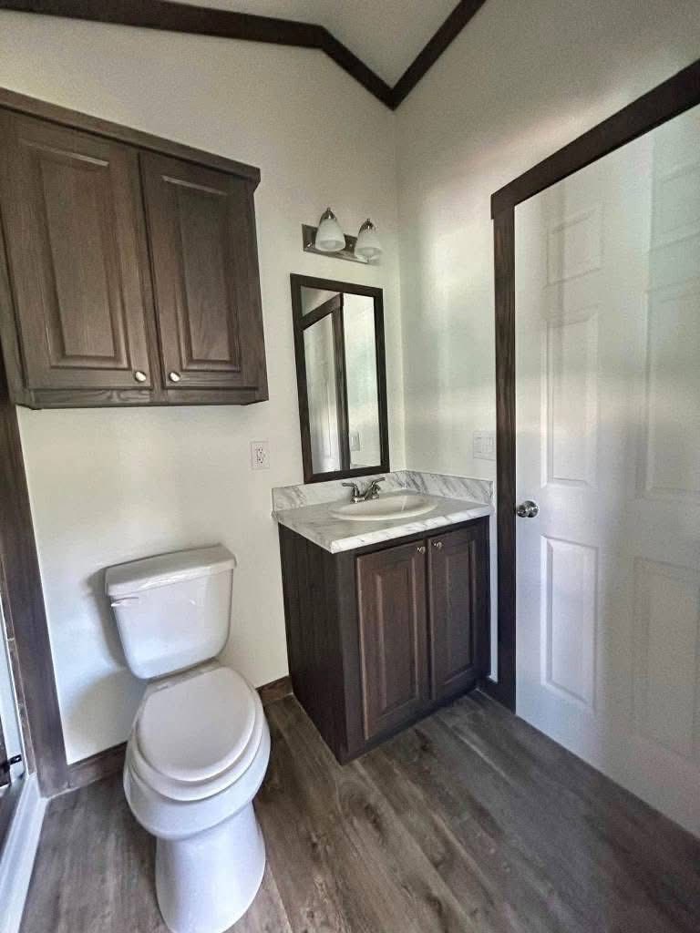 Small bathroom with wood accents. Includes a wooden cabinet above a toilet, a vanity with a mirror and dual lights, and a wooden floor.