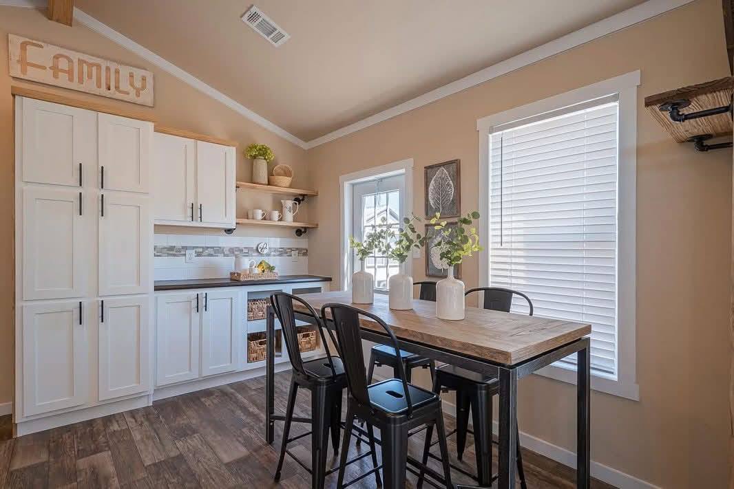 Cozy kitchen with white cabinets and dark countertops, featuring a wooden dining table with black metal chairs. Sunlit window with blinds adds warmth.