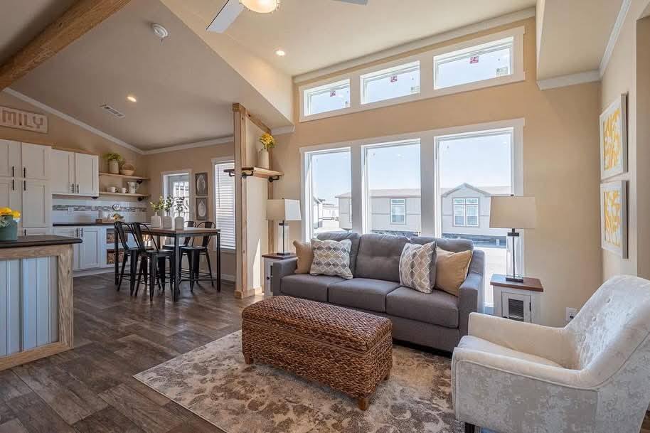 Bright, cozy living room with a large gray sofa, patterned cushions, and a wicker ottoman. Light streams through tall windows, linking to a kitchen.