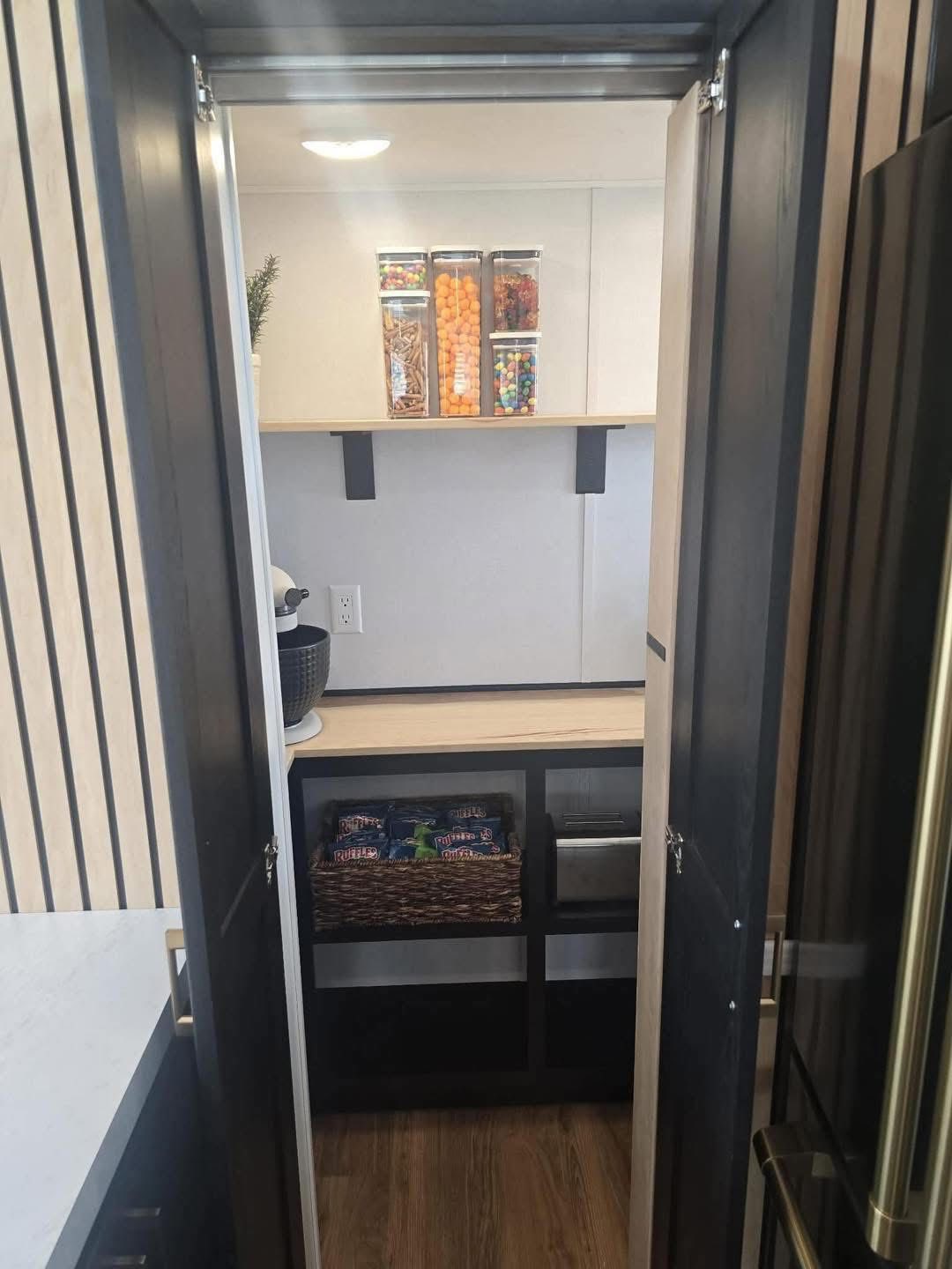 Modern pantry with black shelves and light wood accents. Containers hold colorful snacks on top, with baskets of snacks below, creating an inviting, tidy space.