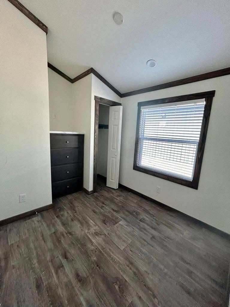 A small, empty room with dark wood floors. It features a window with blinds, white walls, dark wood trim, a black chest of drawers, and an open closet.