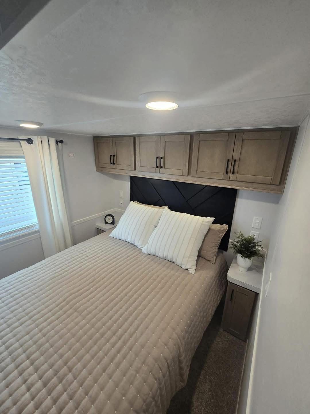 Cozy bedroom with a beige quilted bed, white pillows, and wooden cabinets above. A small clock and plant decorate the bedside, creating a serene atmosphere.