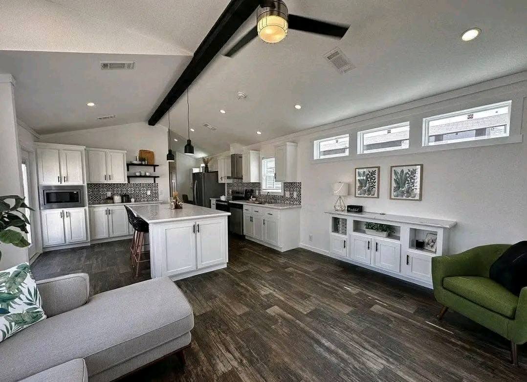 Modern open-concept kitchen and living room with white cabinets, dark wood floors, and black ceiling beam. Cozy green chair and decorative plants accentuate the space.