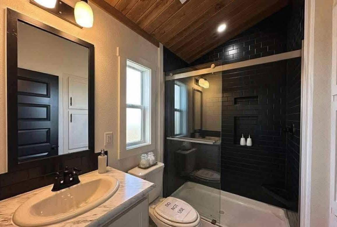 Modern bathroom featuring a sloped wooden ceiling, white sink with a marble countertop, black tiled shower, and a "Do Not Use" sign on the toilet.