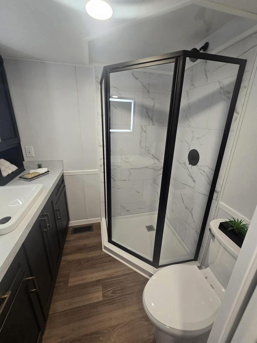 Modern bathroom with wood floor, featuring a marble-tiled shower with a glass door. Vanity with sink on the left, and toilet on the right. Bright and clean.