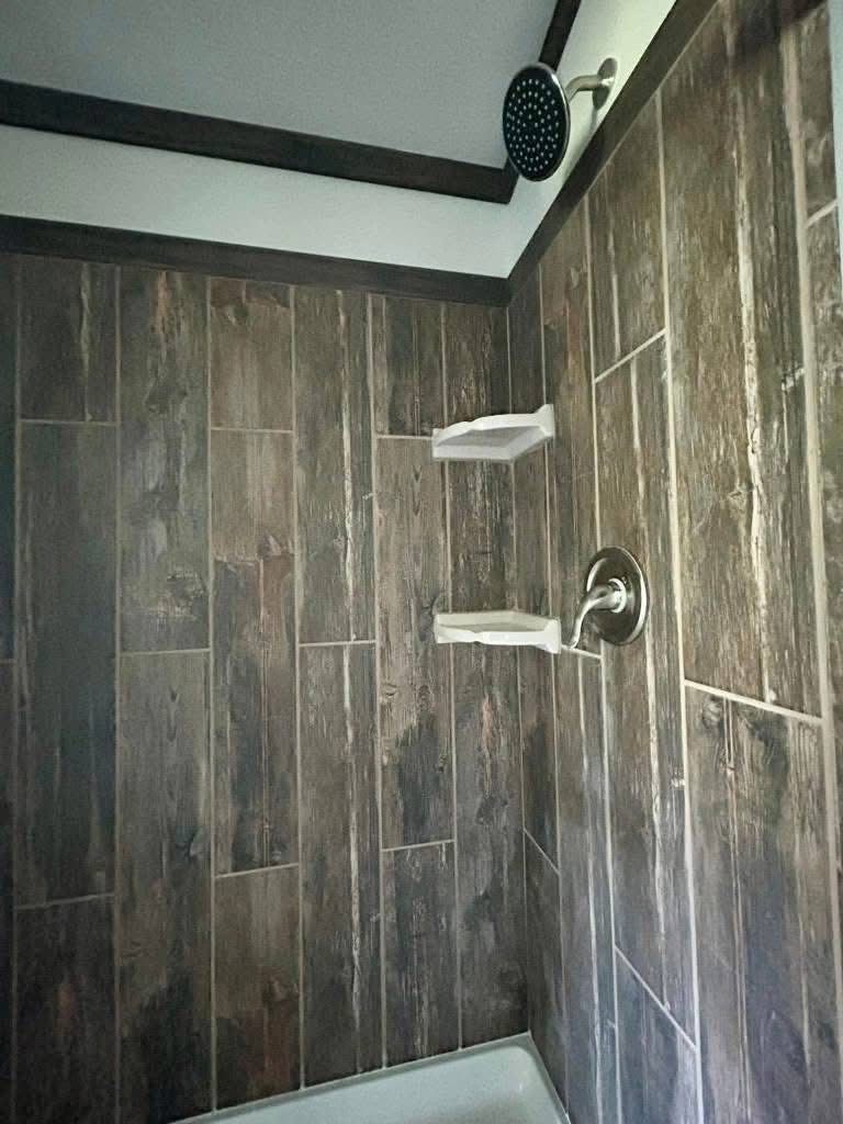 Shower interior with wood-textured tiles, two white corner shelves, and a black round showerhead, creating a rustic and modern ambiance.