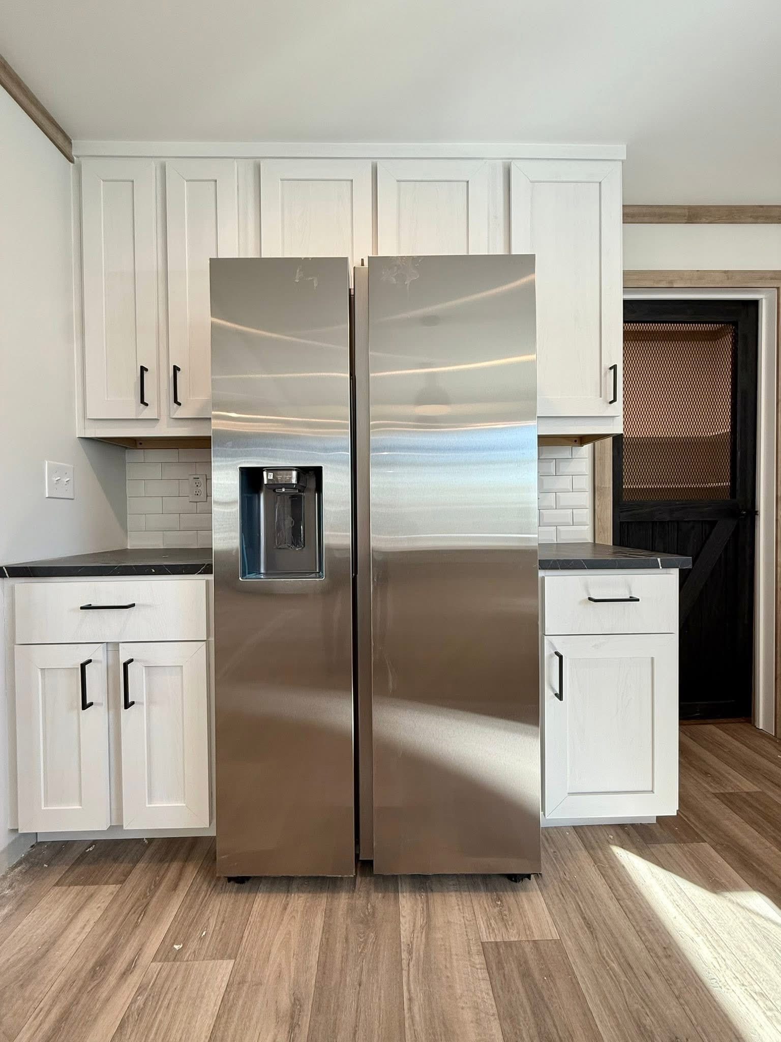 A sleek stainless steel refrigerator stands centered in a kitchen with white cabinets and a dark countertop. Light wood flooring complements the modern design.