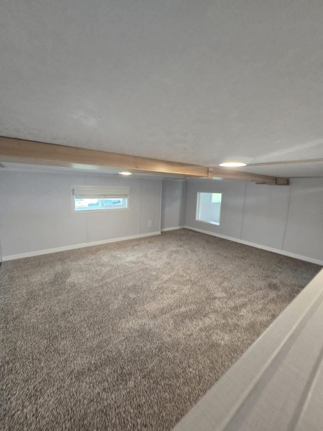 A small, cozy attic room with gray carpet, two small windows, and exposed wooden beams. The space is well-lit, creating a serene atmosphere.