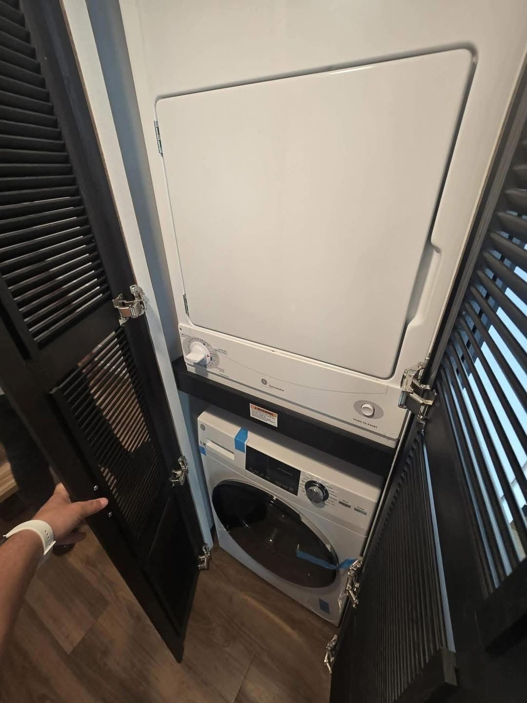Stacked washer and dryer inside a narrow closet with dark louvered doors. A person’s hand is opening the door, revealing the compact appliances.