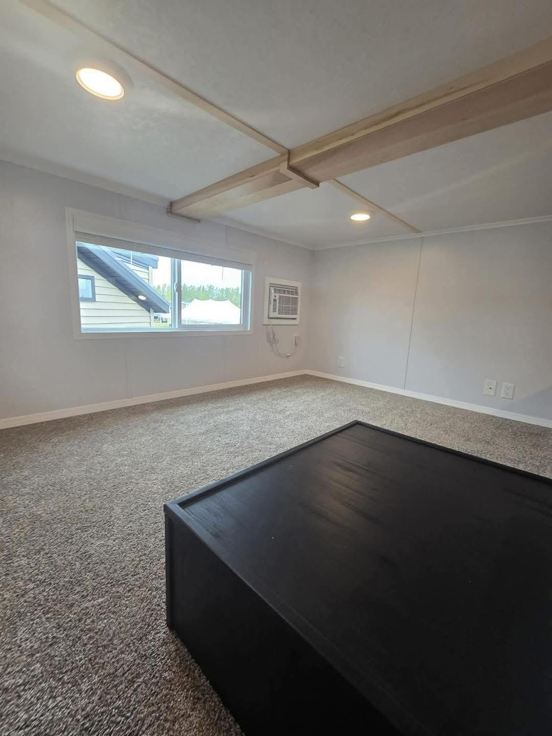 Small, bright room with grey carpet and white walls. Large window and air conditioner on right wall. Black square table in foreground. Modern, minimalistic feel.