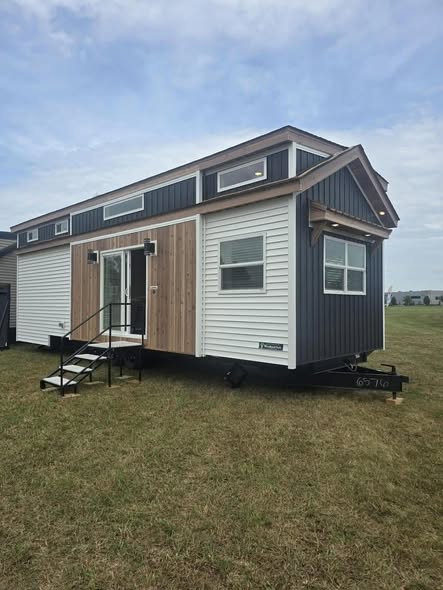 A modern tiny house with contrasting wood and dark siding sits on a grassy field under a partly cloudy sky, exuding a minimalist and cozy vibe.