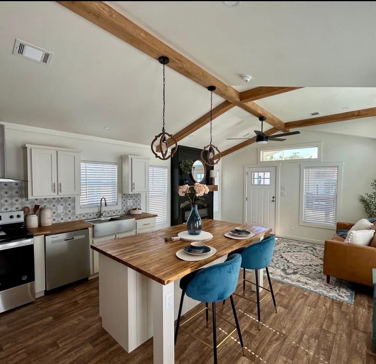 Modern kitchen-living room with wooden beams, island bar with blue stools, pendant lights, white cabinets, and cozy seating area. Bright, inviting space.