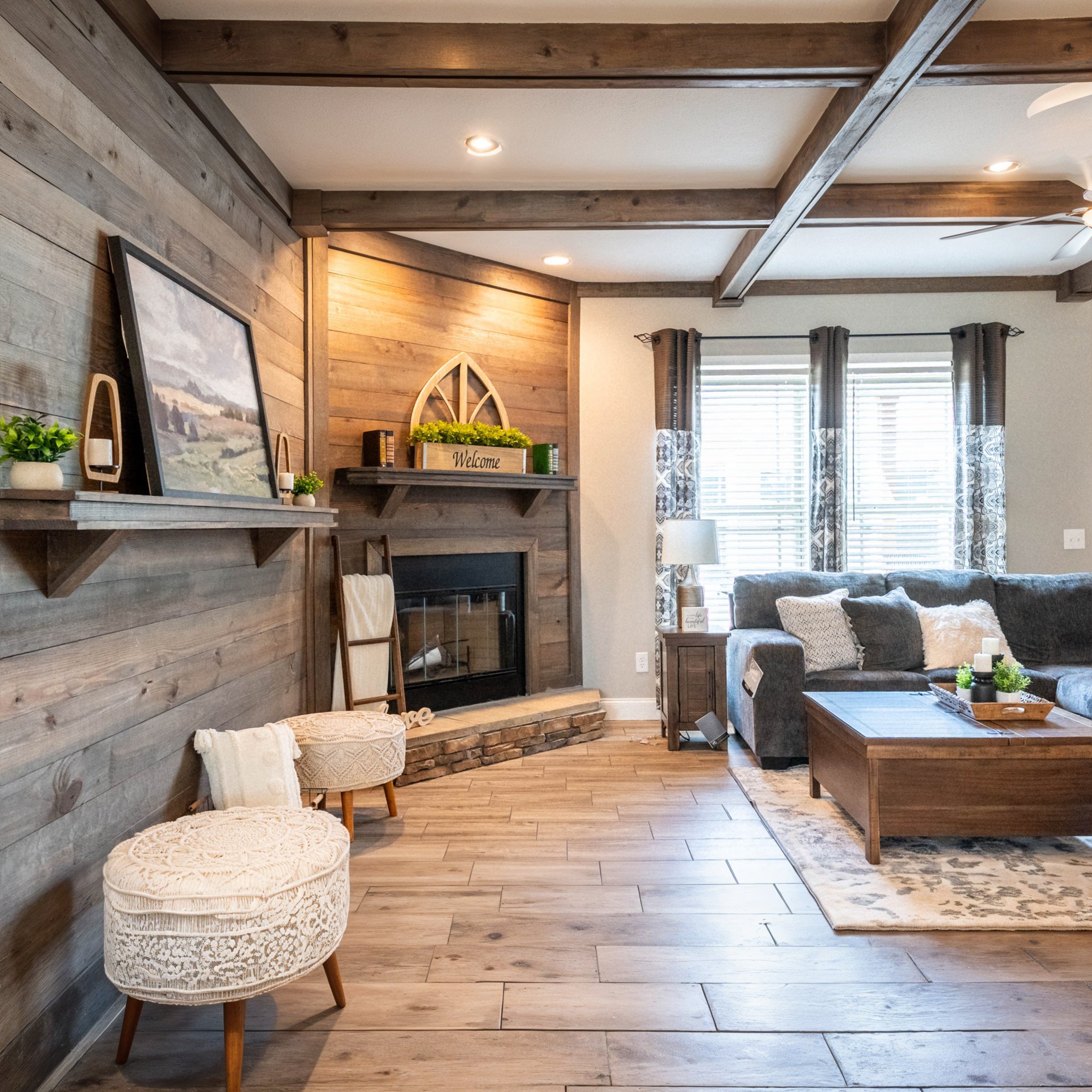 Cozy living room with wood-paneled walls, rustic fireplace, and beamed ceiling. Features a comfortable gray sofa, soft cushions, and welcoming decor.