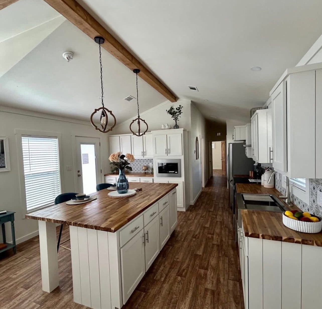 A modern kitchen with white cabinets and wooden countertops. Two pendant lights hang above a central island with a vase and flowers. The room is bright and clean.